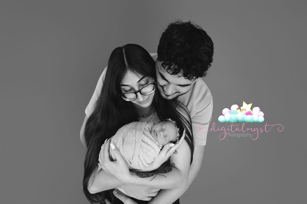 Black and white photo of parents lovingly cradling their newborn baby against a gray studio backdrop.
