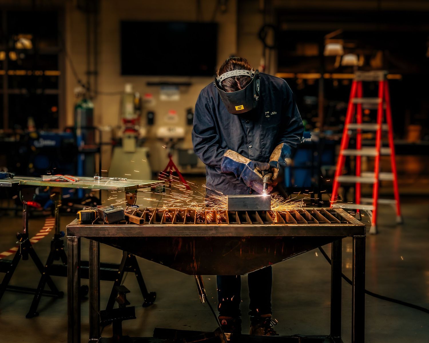 Welder in protective gear creates sparks while welding metal on a workbench in an industrial workshop.