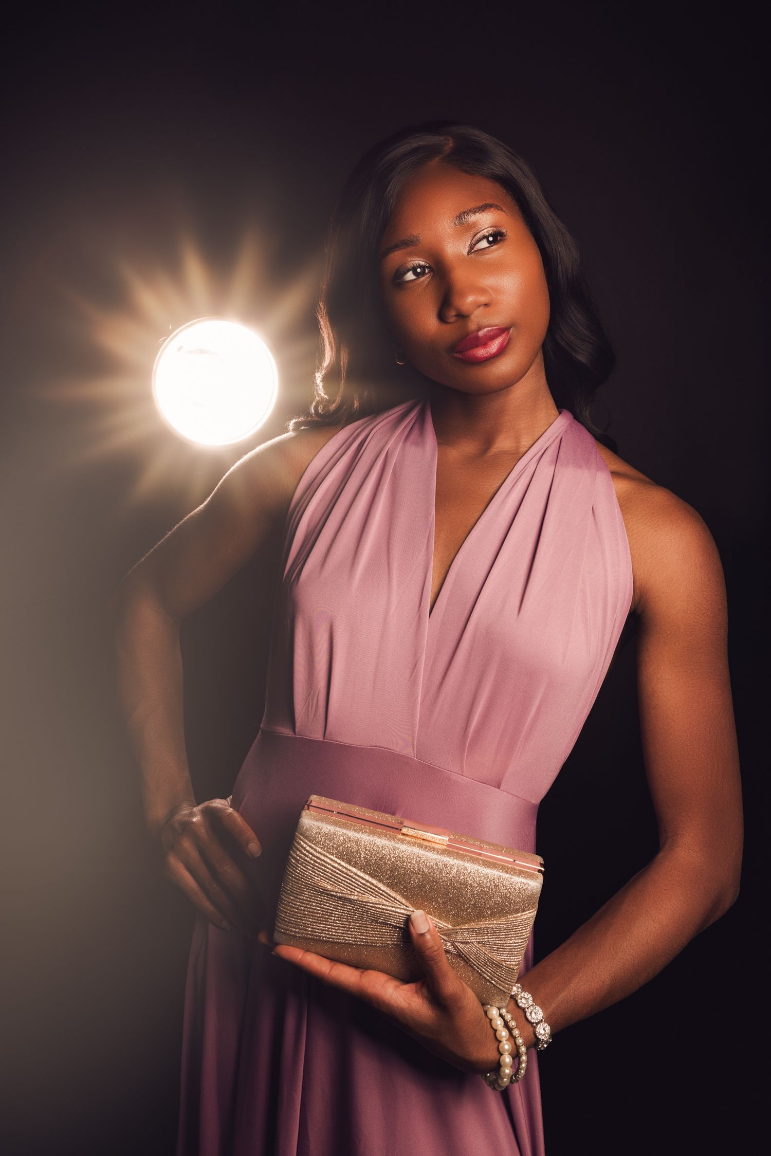 Elegant Black woman in pink halter gown holding gold clutch, posed before bright studio light on dark background.