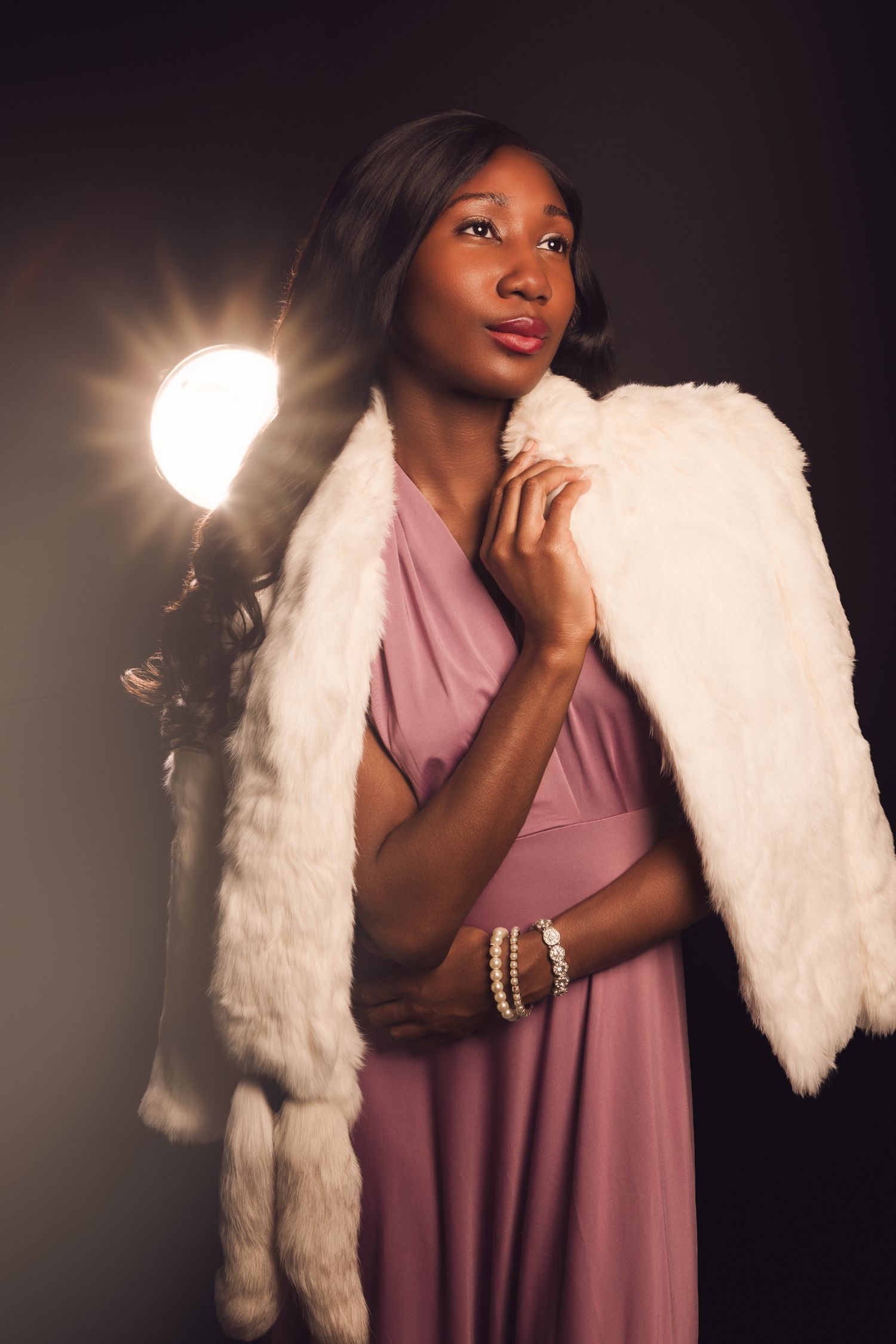 Elegant Black woman in pink gown and white fur stole poses gracefully before a studio light on dark background.