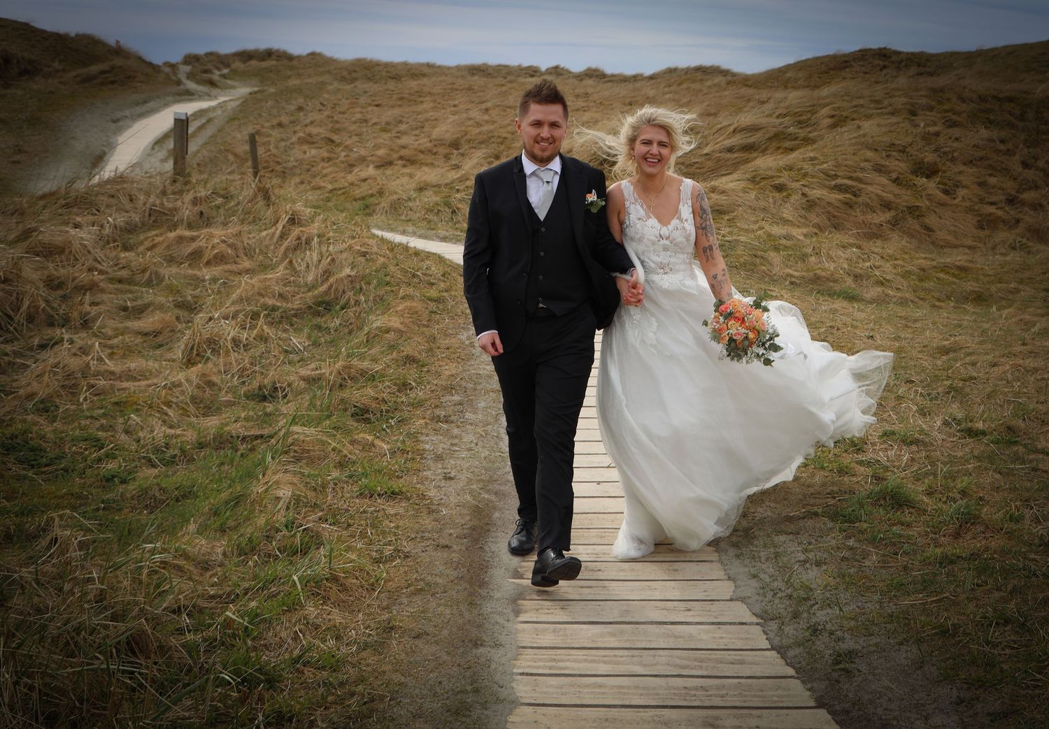 Bride and groom walk hand in hand along wooden boardwalk through coastal dunes on their wedding day.