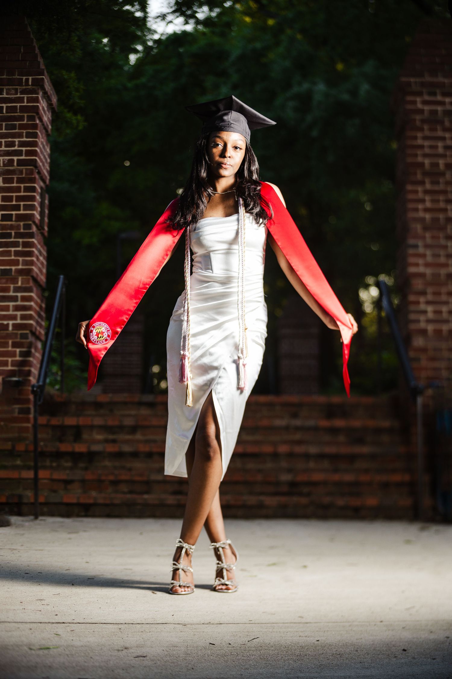 Young woman in white dress and graduation cap poses proudly with red sash outdoors near brick wall.