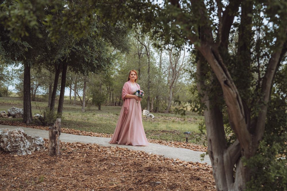 Woman in elegant pink gown holding bouquet stands on garden path surrounded by lush green trees and autumn leaves.