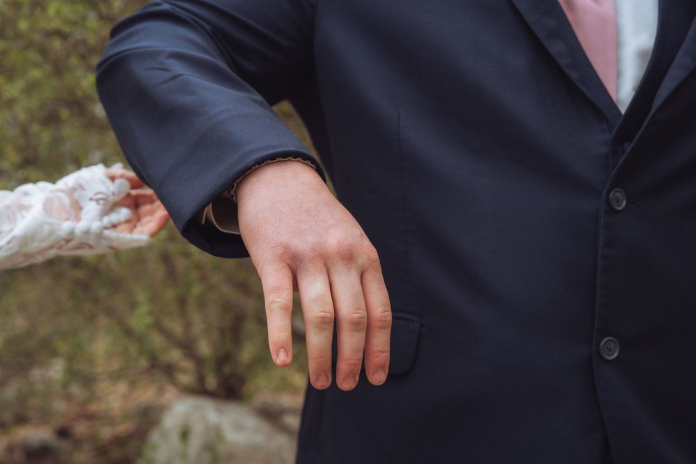 Close-up of groom's hand wearing wedding ring in navy suit with pink tie outdoors.