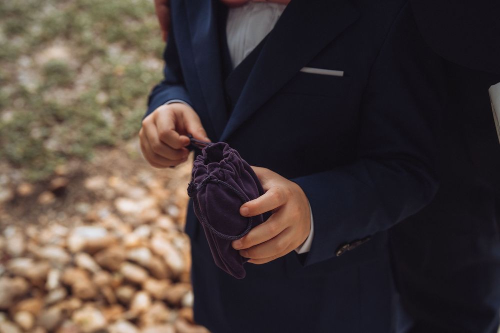 Man in navy suit holding a small purple drawstring pouch outdoors with autumn leaves in background.