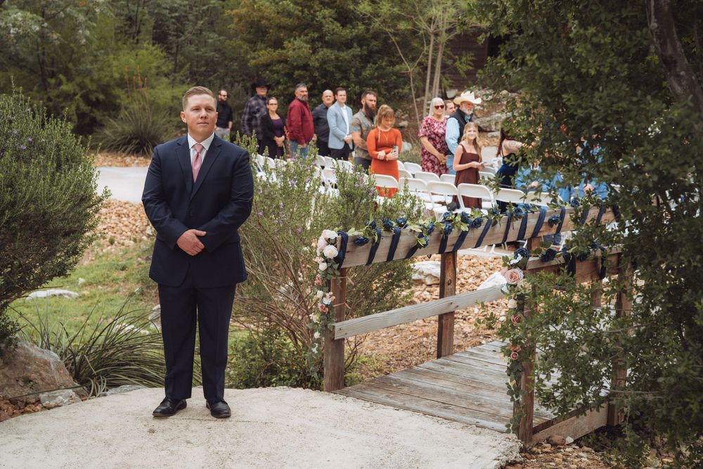 Man in dark suit stands near wooden bridge at outdoor wedding ceremony with guests gathered in background.