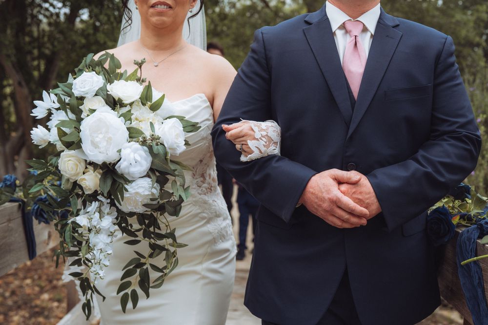 Bride holding white peony bouquet walks arm in arm with suited escort at wedding.