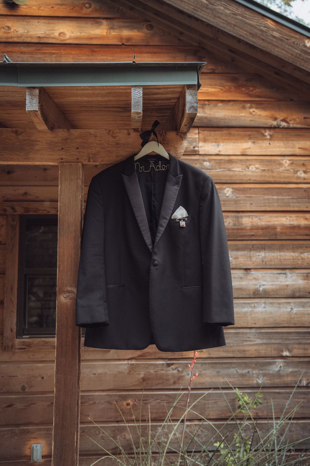 Black tuxedo jacket with boutonniere on wooden hanger displayed outside rustic log cabin wedding venue.