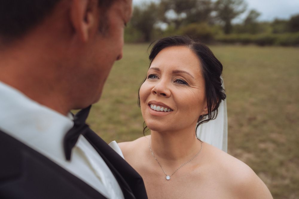 Bride smiling at groom during outdoor wedding ceremony in green field, wearing strapless gown and veil.