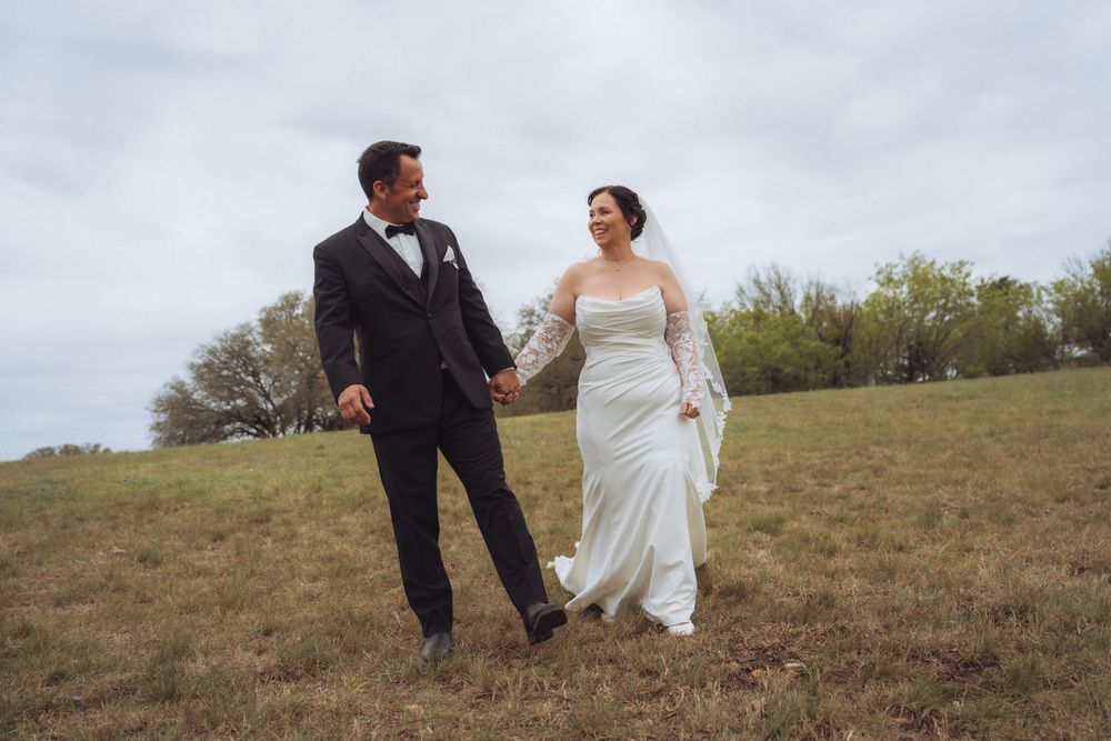 A bride and groom hold hands while walking through a green field on their wedding day under a cloudy sky.
