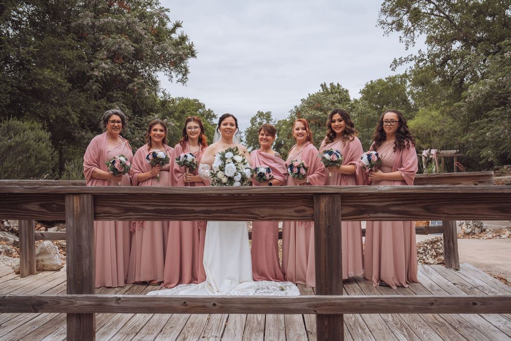 Bride and bridesmaids in pink dresses posing on a wooden bridge surrounded by trees.