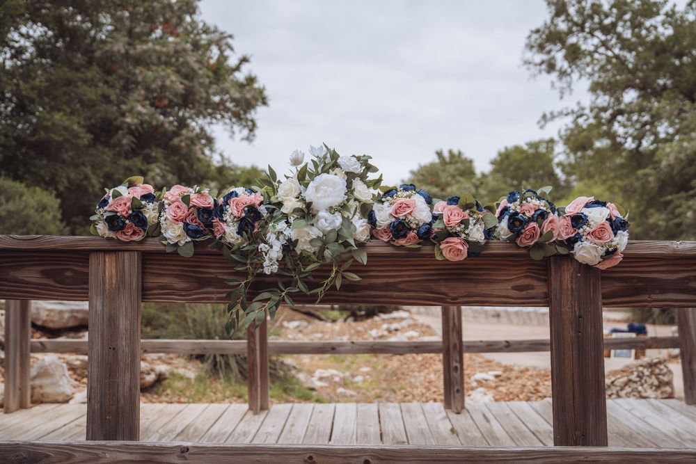 Pink, white, and navy wedding bouquets arranged on rustic wooden bridge railing at outdoor ceremony.