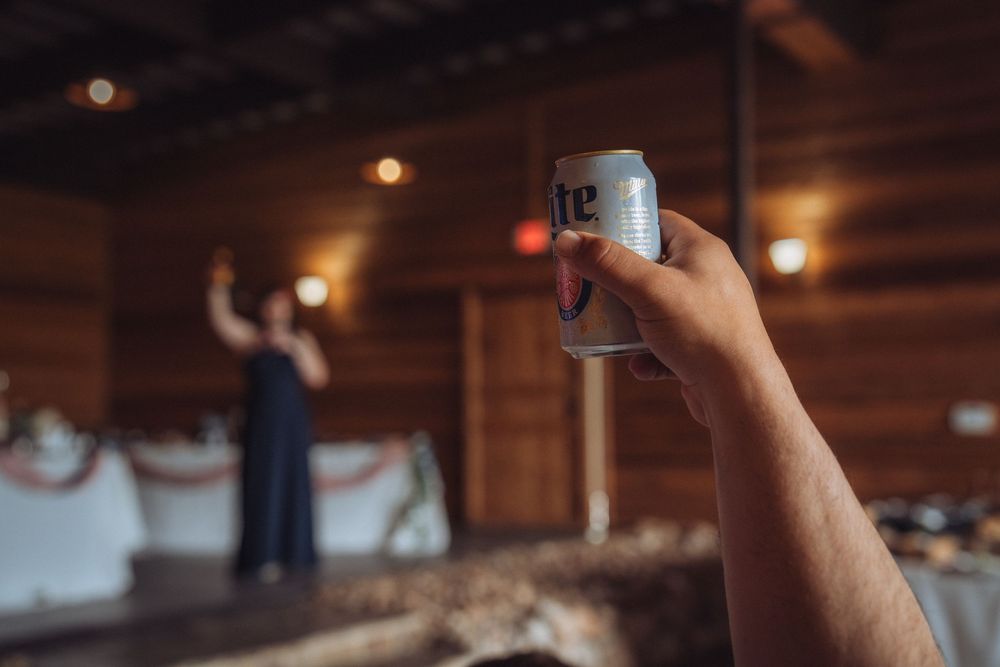 Guest holding Miller Lite beer can at rustic barn wedding reception with bridesmaid dancing in background.