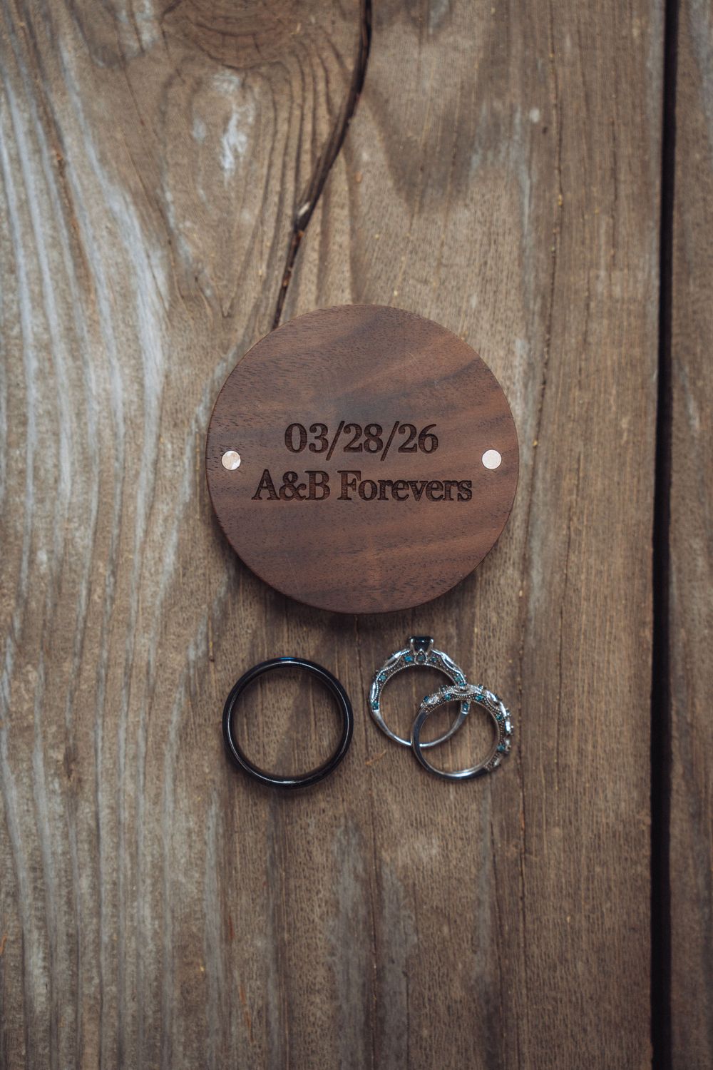 Wooden wedding ring holder engraved with 03/28/26 A&B Forevers beside two wedding rings on rustic wood.