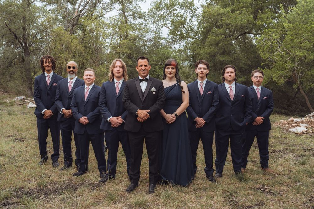 Wedding party in navy suits and black gown posing outdoors in a grassy field with trees in the background.
