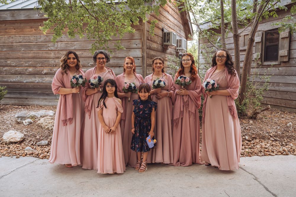 Bridesmaids in dusty rose gowns pose with two flower girls in front of a rustic wooden barn.