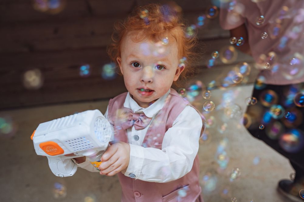 Adorable redhead toddler in a vest holding a bubble gun at a wedding celebration.