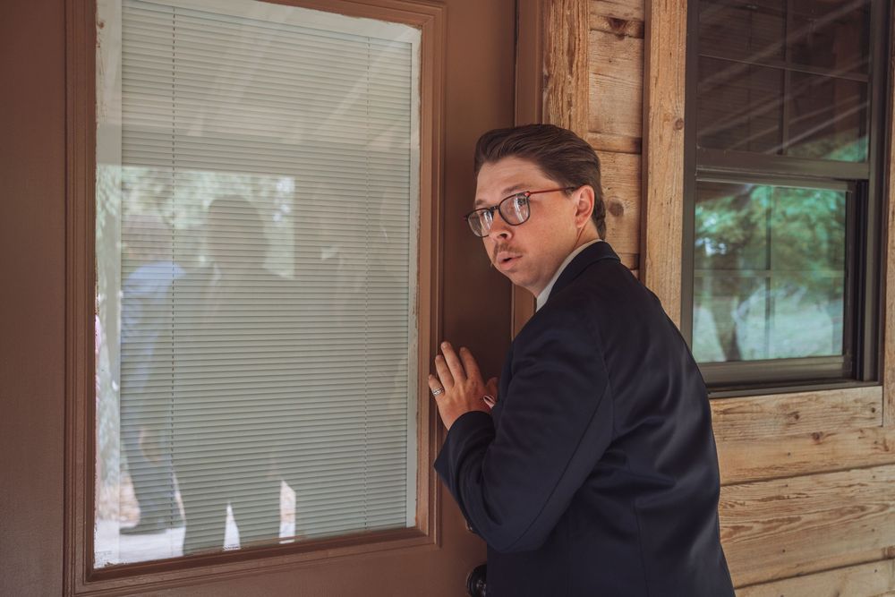 Man in suit pressing against wall peering through window blinds in wooden building.