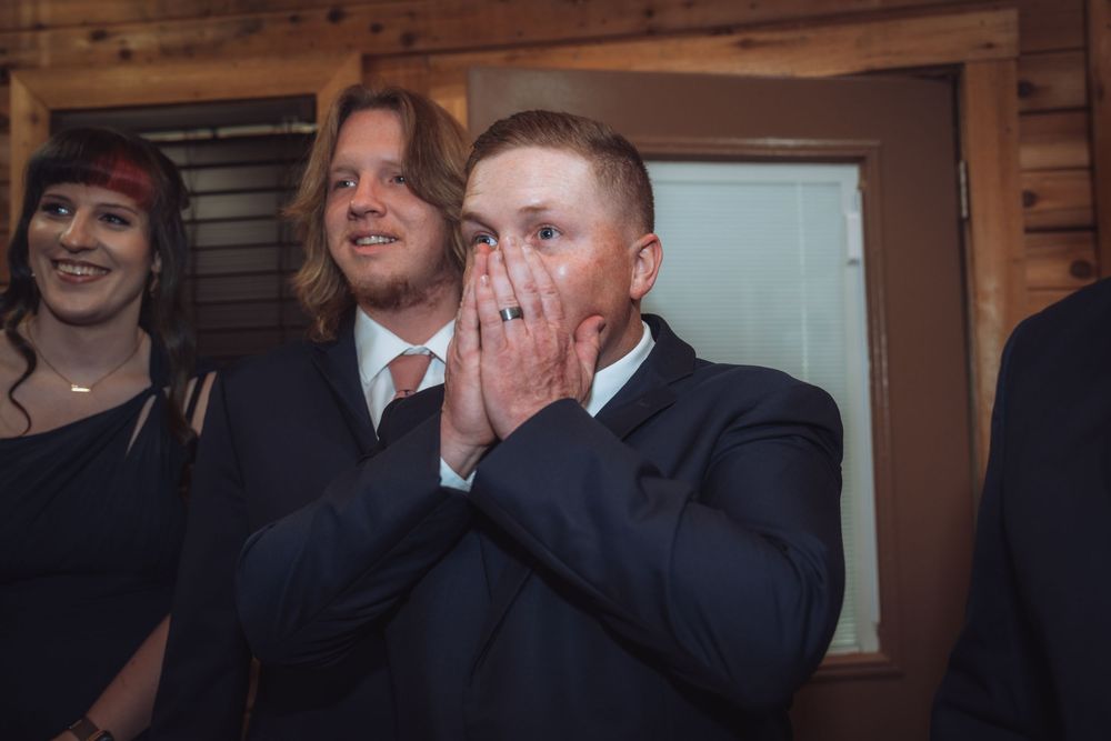 Emotional groom in navy suit covering mouth with hands at wedding ceremony, surrounded by guests in rustic venue.