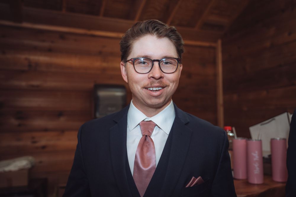 Smiling groom in navy suit with pink tie and pocket square wearing glasses in rustic wood cabin setting.