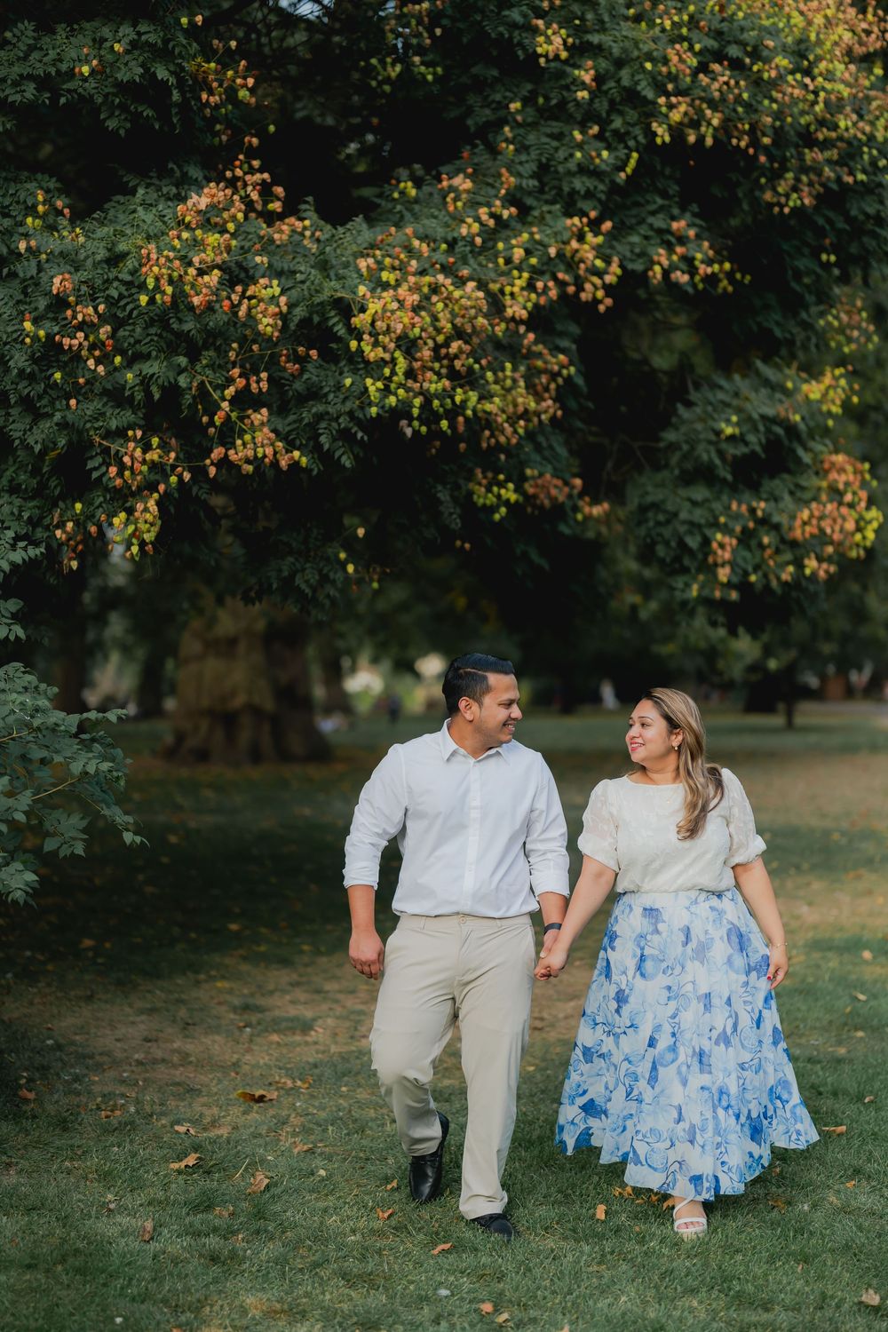 A couple walks hand-in-hand through a lush green park, smiling at each other, dressed in casual elegant attire.