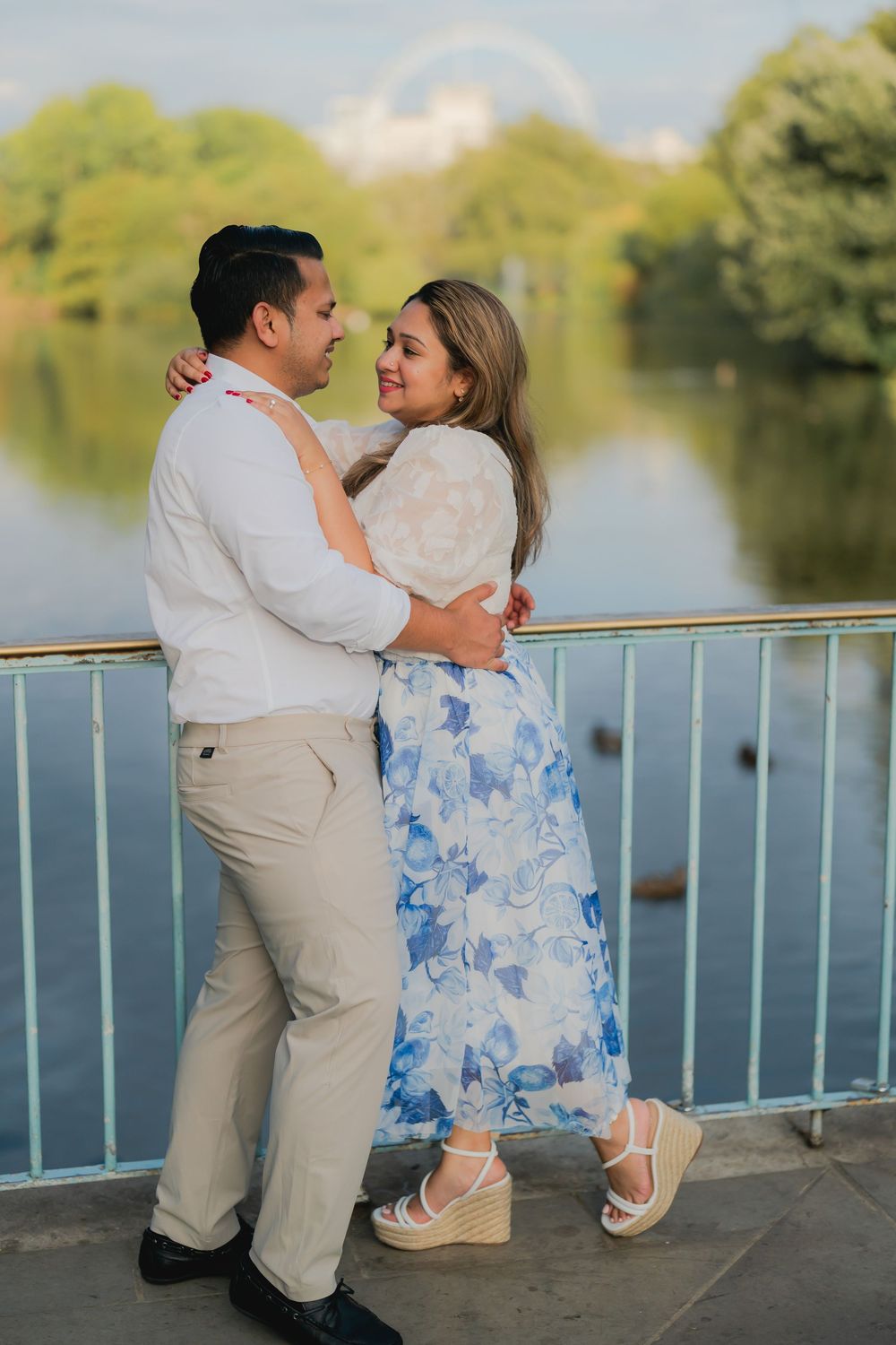 Couple embracing on a bridge by a serene lake, woman in blue floral dress and man in white shirt, sharing a romantic moment.