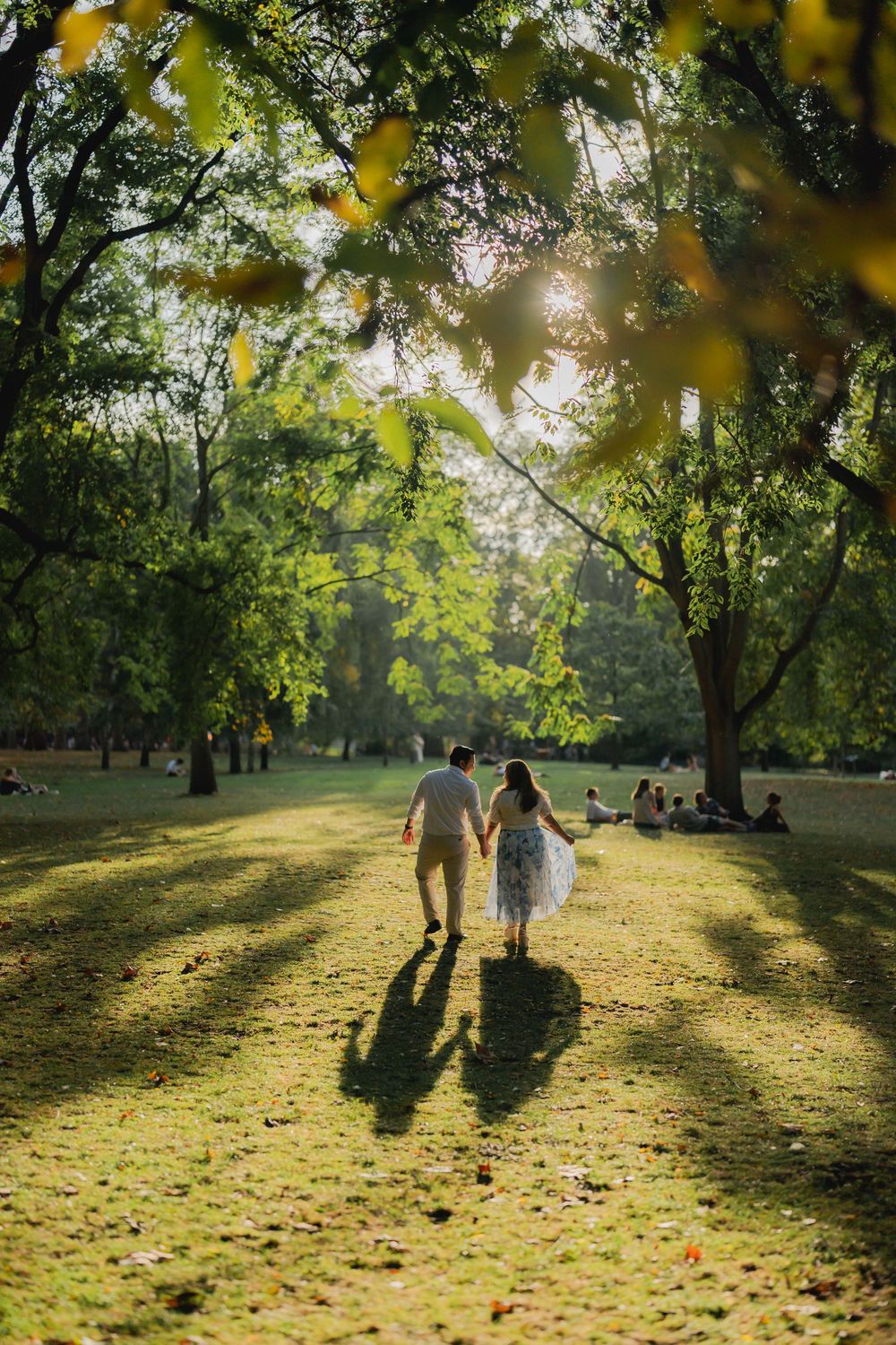 Couple holding hands walking through a sunlit park with lush green trees and golden light at sunset.