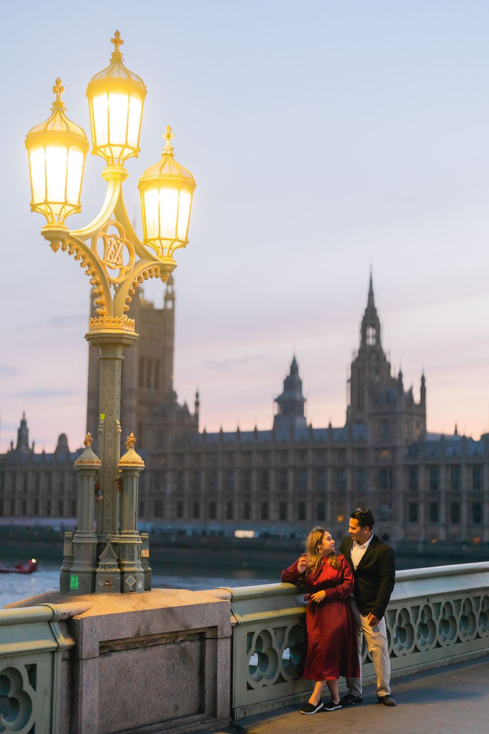Couple standing by a glowing lamppost on Westminster Bridge with Houses of Parliament at dusk.