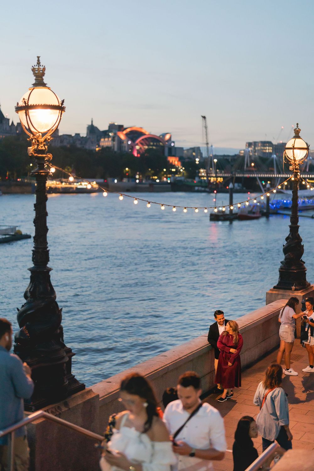 People strolling along a riverside promenade at dusk, illuminated by ornate street lamps with city lights reflected on water.
