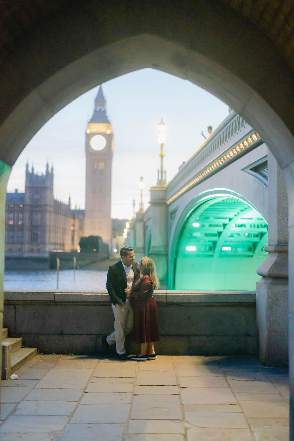 Couple sharing a romantic moment under an arch near Westminster Bridge with Big Ben illuminated at dusk in London.