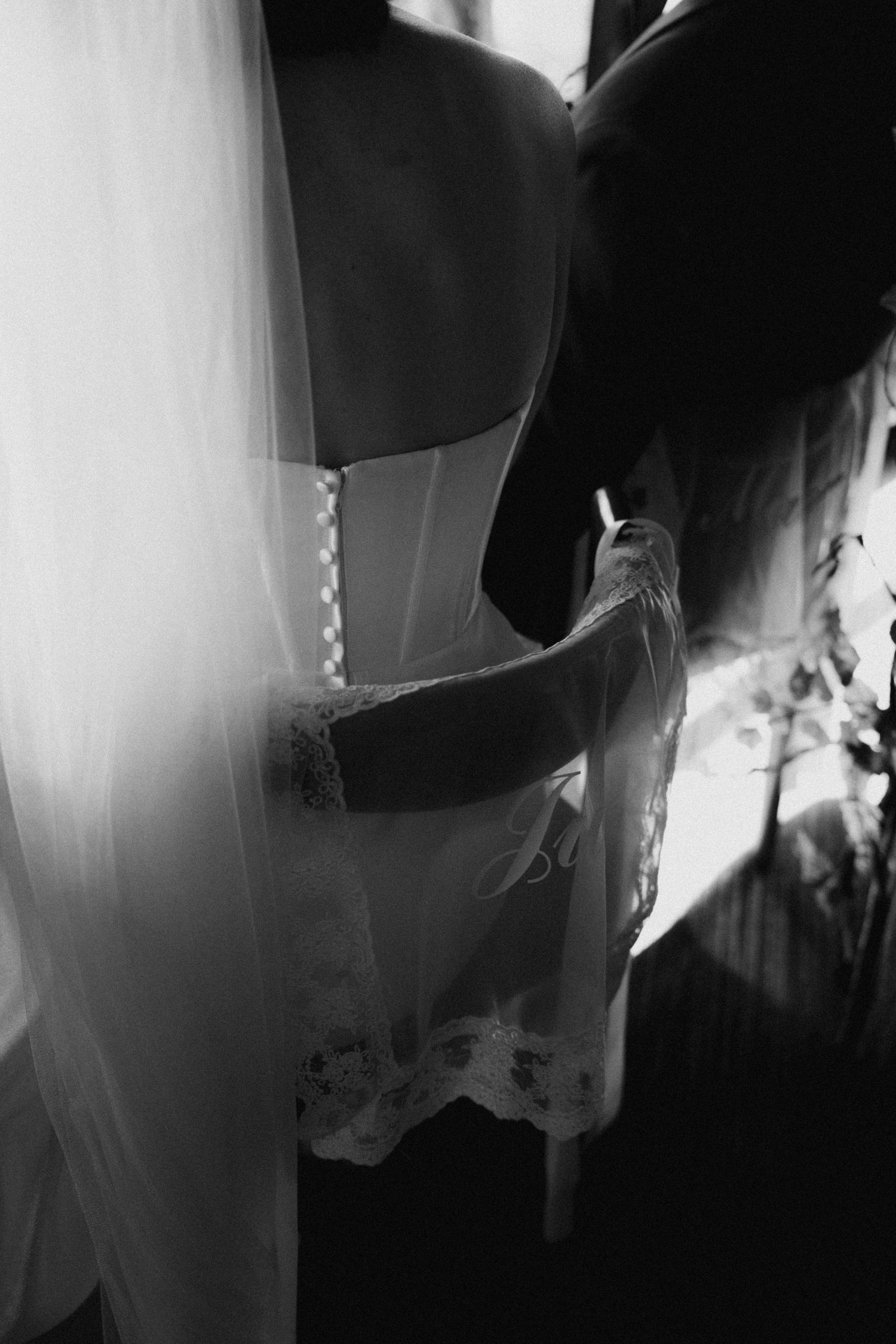 Black and white photo of bride getting corset wedding dress laced up by bridesmaid near window with veil.