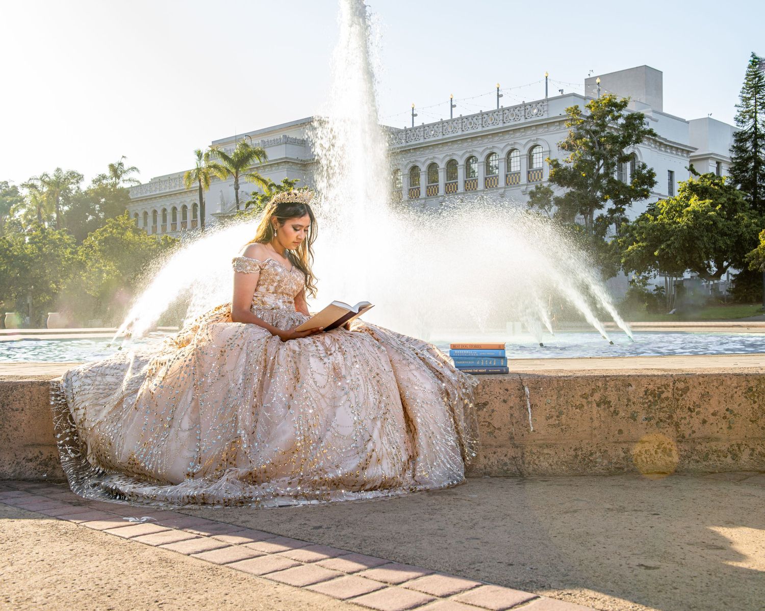 Young woman in elegant quinceañera gown reading by a fountain, with a grand white building backdrop in sunlight.