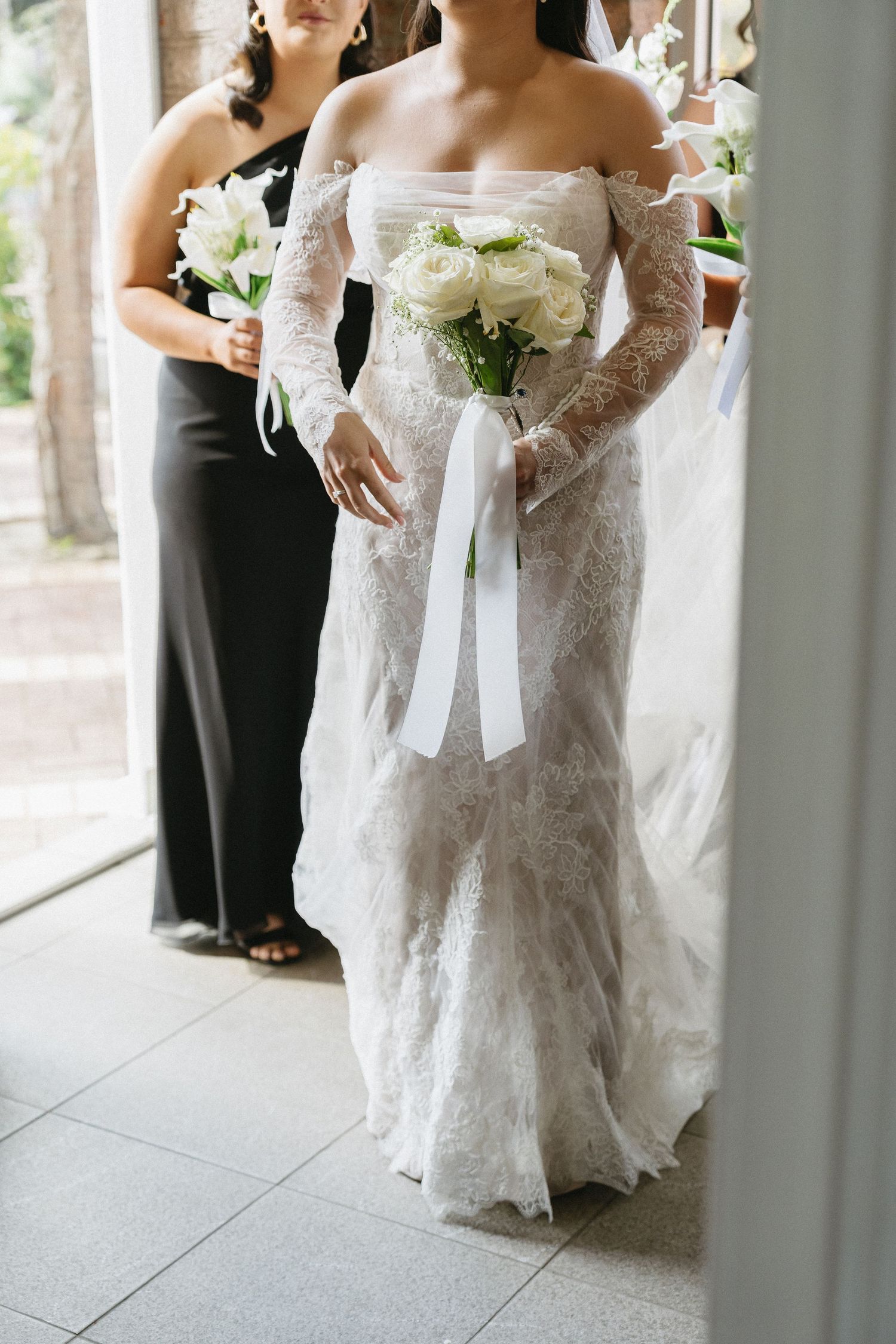 Bride in off-shoulder lace wedding gown holding white rose bouquet with ribbon, bridesmaid in black dress behind her.