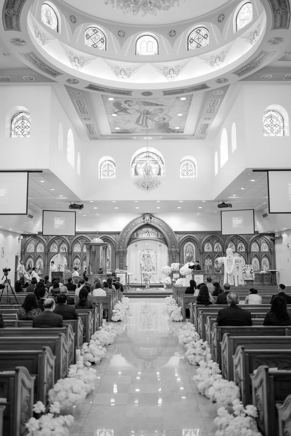 Black and white photo of elegant church wedding ceremony with floral aisle decorations and ornate domed ceiling.