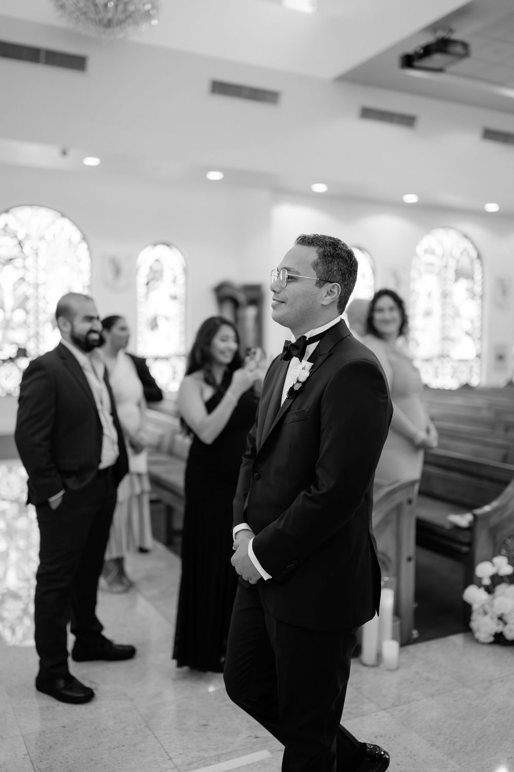 Groom in black tuxedo smiles with anticipation at the altar during church wedding ceremony.