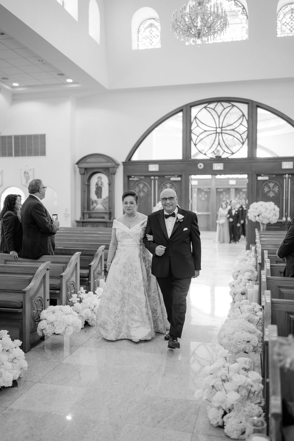 Bride walks down flower-lined church aisle escorted by her father in a black tuxedo.