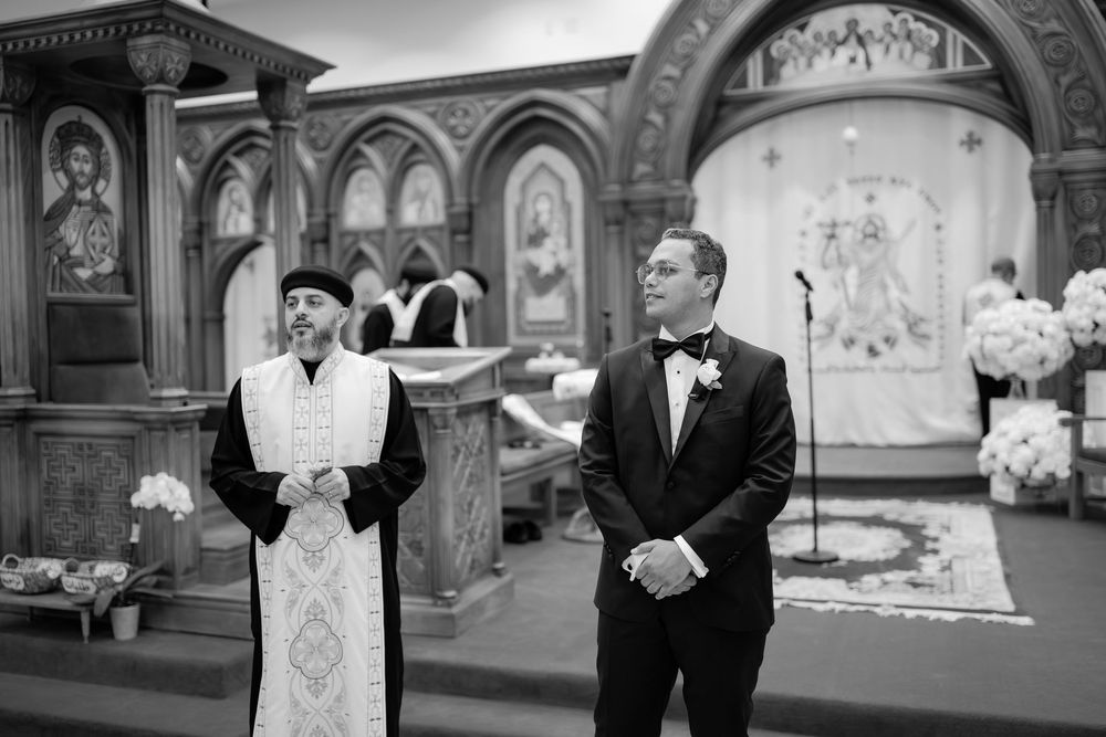 Groom in tuxedo stands with priest in ornate church during wedding ceremony.