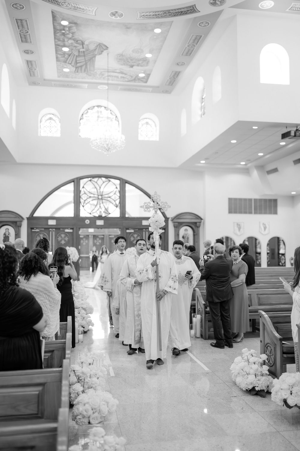 Black and white photo of clergy processing down a flower-lined church aisle during a wedding ceremony.