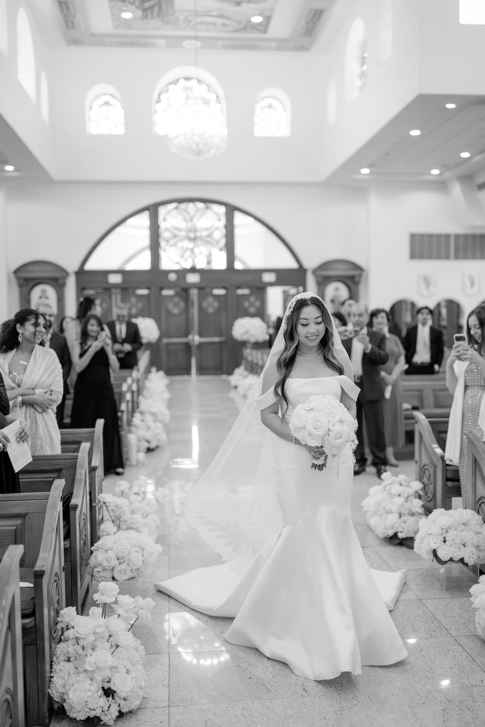 Bride in off-shoulder mermaid gown walks down church aisle holding bouquet, guests watching, black and white wedding photo.