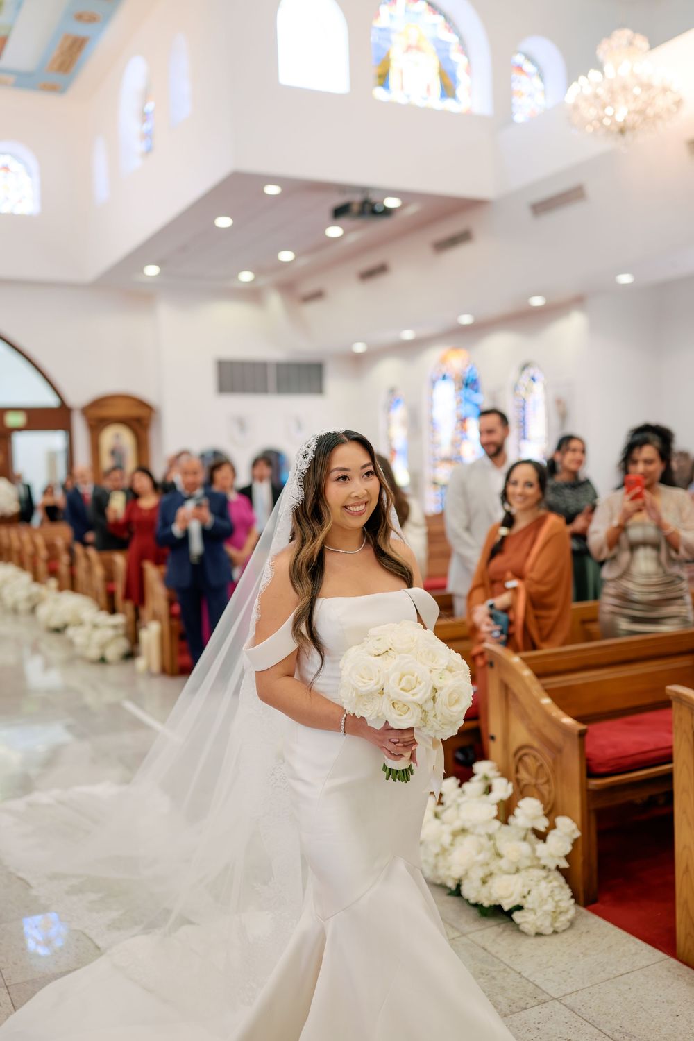 Black and white photo of a bride walking down the church aisle in a white gown, holding a bouquet, with guests seated in pews.