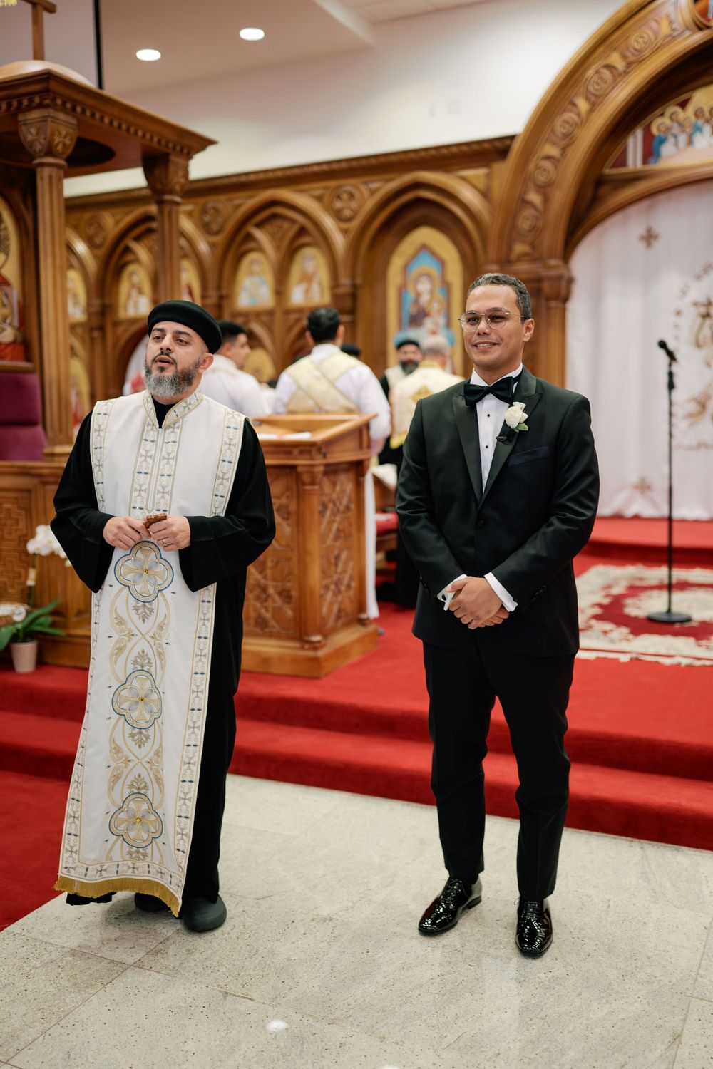 Groom in tuxedo stands with priest in ornate robes at church altar during wedding ceremony, black and white photo.