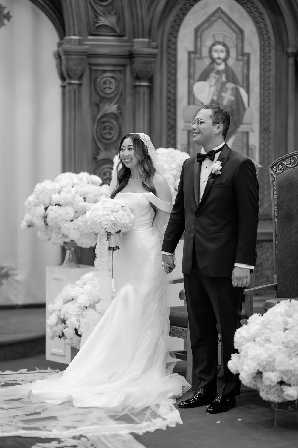 Black and white photo of a bride and groom standing at the altar of a church decorated with white floral arrangements.