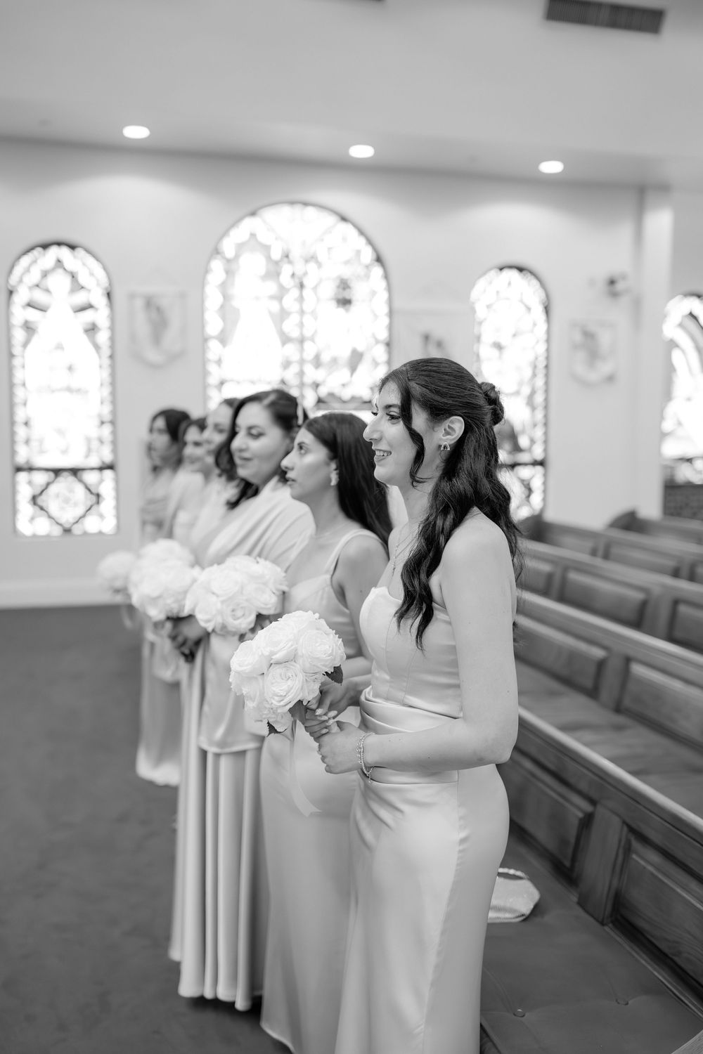 Bridesmaids in elegant gowns holding white bouquets stand in a church aisle in black and white.