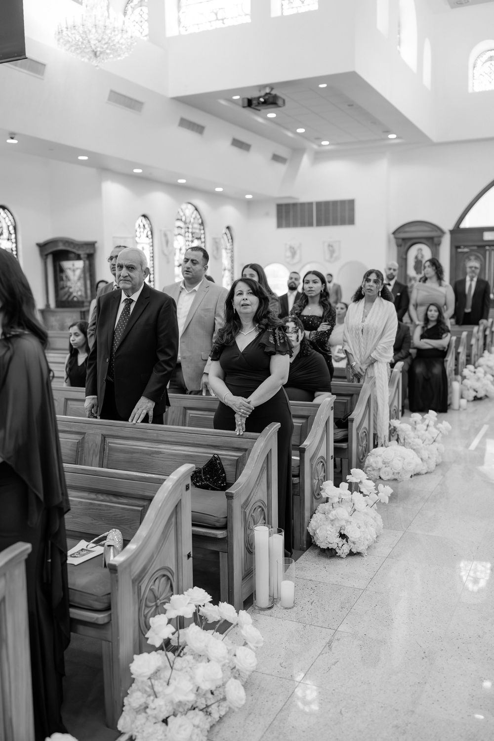 Guests stand in church pews during a wedding ceremony, with floral arrangements and candles lining the aisle.