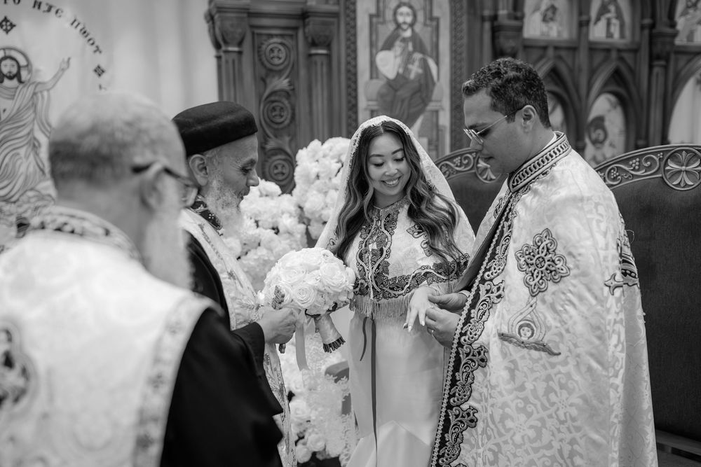 Black and white Orthodox wedding ceremony photo with bride holding bouquet, groom, and priest at ornate church altar.