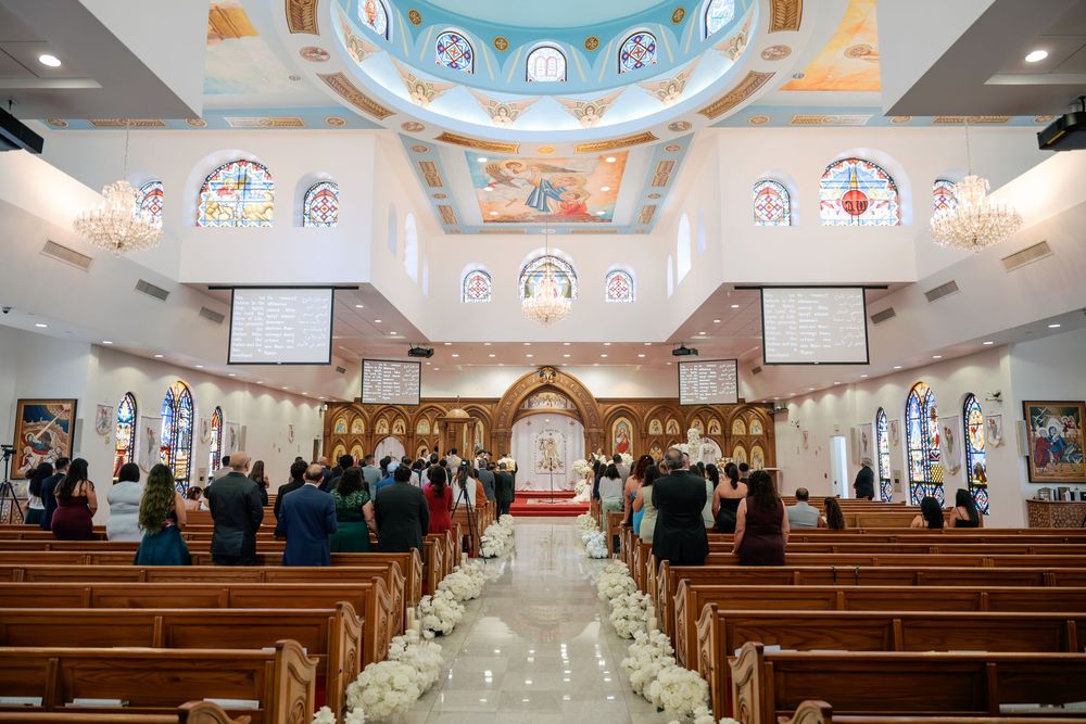 Black and white wedding ceremony photo inside an elegant church with floral aisle decorations and guests seated in pews.