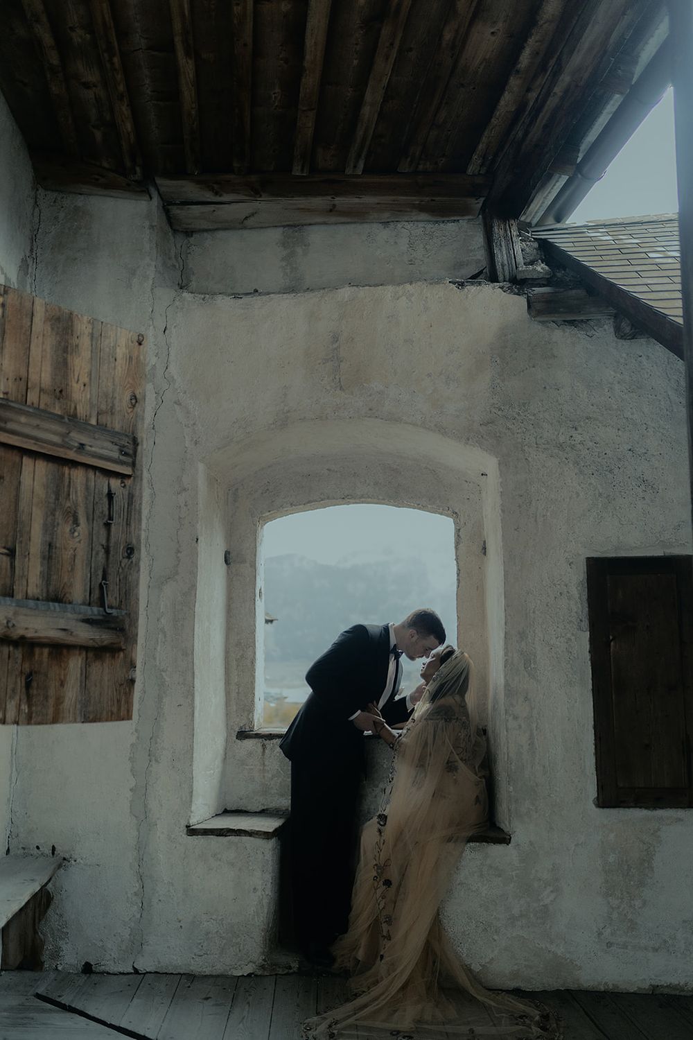 Wedding couple kissing by arched stone window with mountain views in rustic historic building.