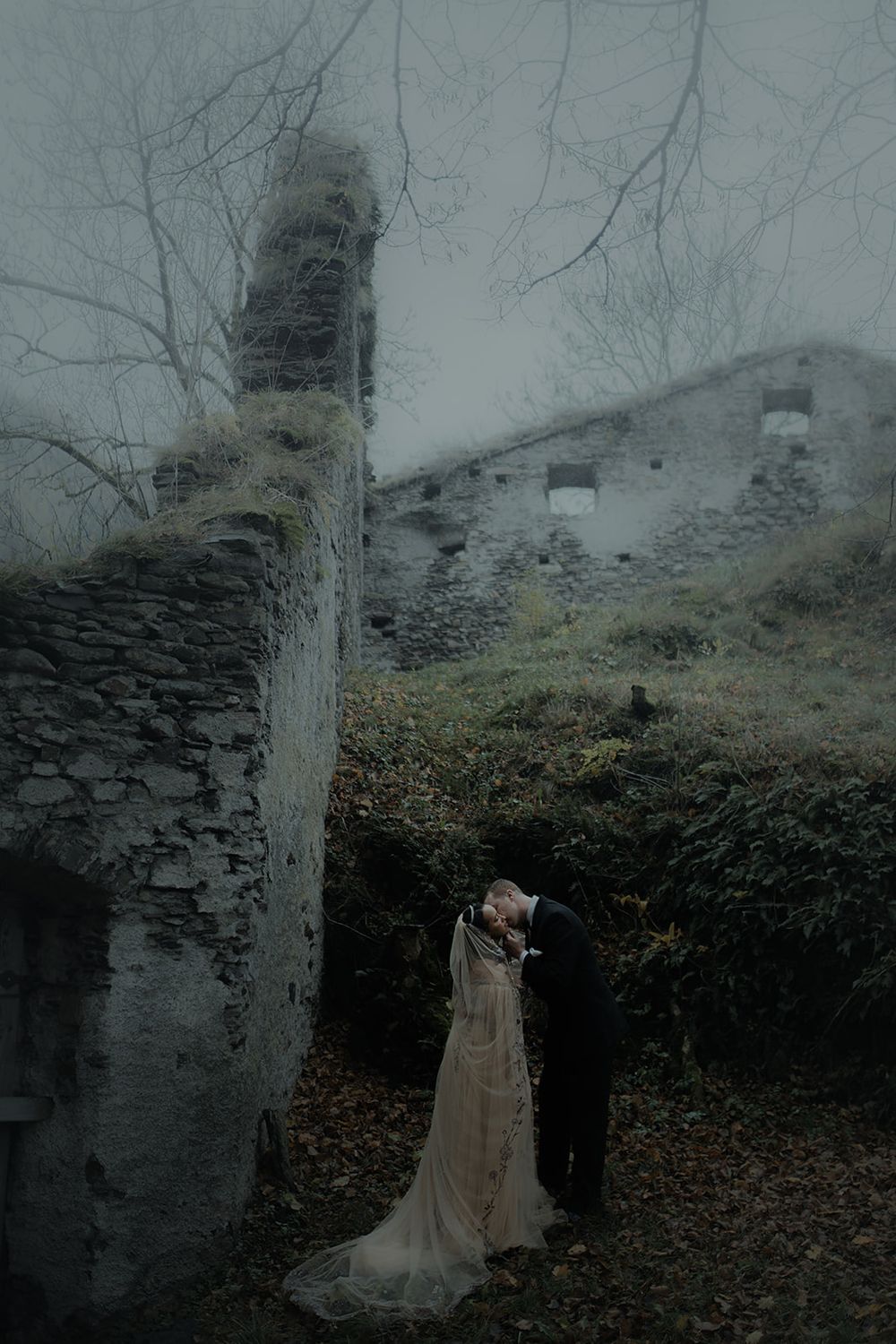 Couple in wedding attire kissing among misty ruins and bare trees in a moody, atmospheric outdoor setting.