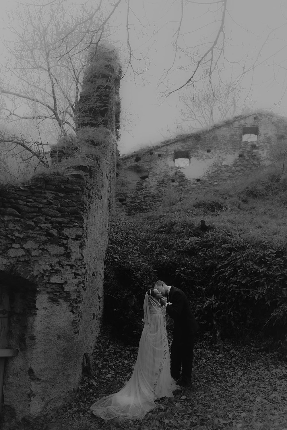 Black and white photo of wedding couple embracing near ancient stone ruins on a foggy, overcast day.