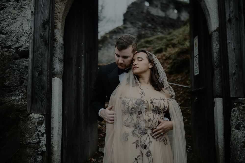 Romantic couple embracing in gothic stone archway, bride in floral gown with veil, groom in black suit.